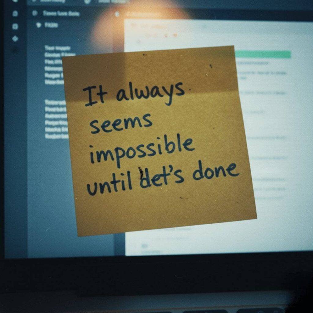 "It always seems impossible until it's done" written on a grainy sticky note stuck to a dimly lit laptop screen.