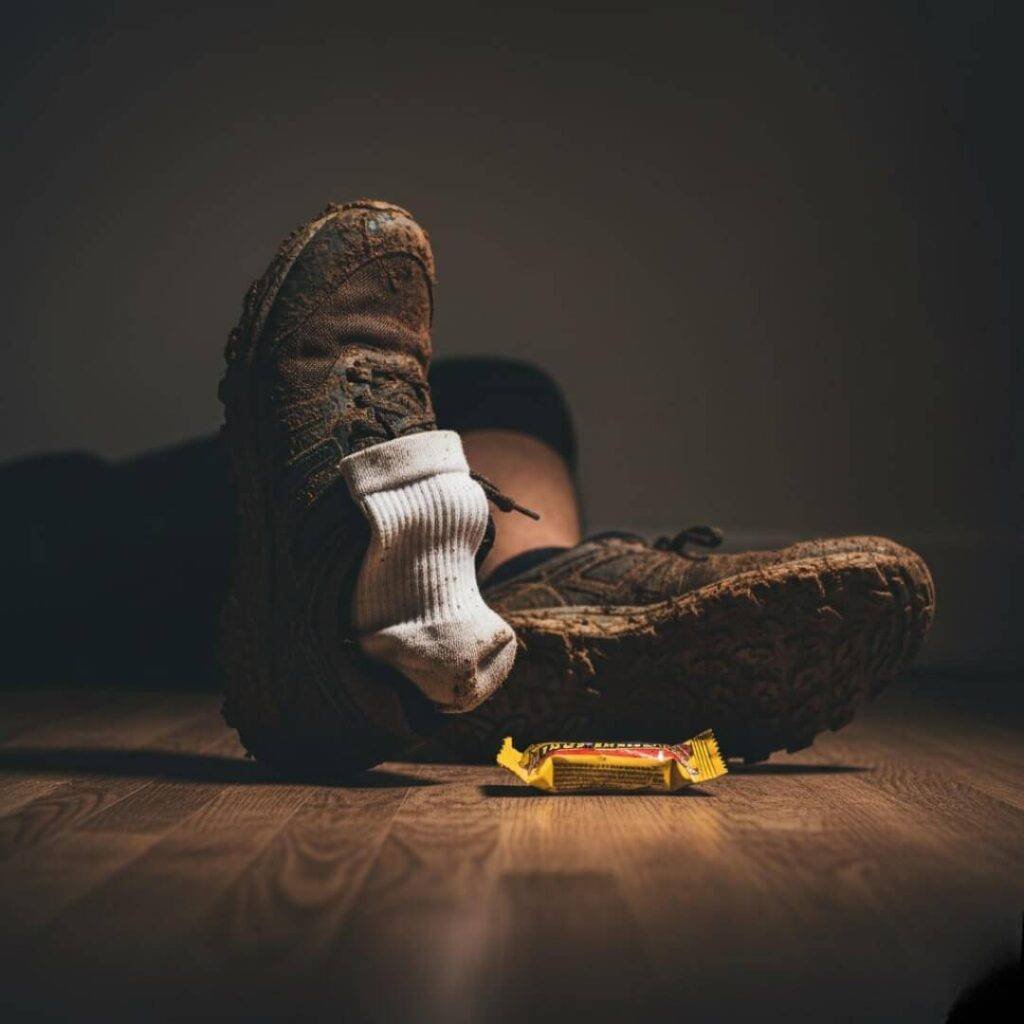 Alt Text: A dim, low-angle close-up of a pair of muddy running shoes on a floor, next to a crumpled energy bar wrapper. A dirty white sock is haphazardly stuck to the side of one shoe.