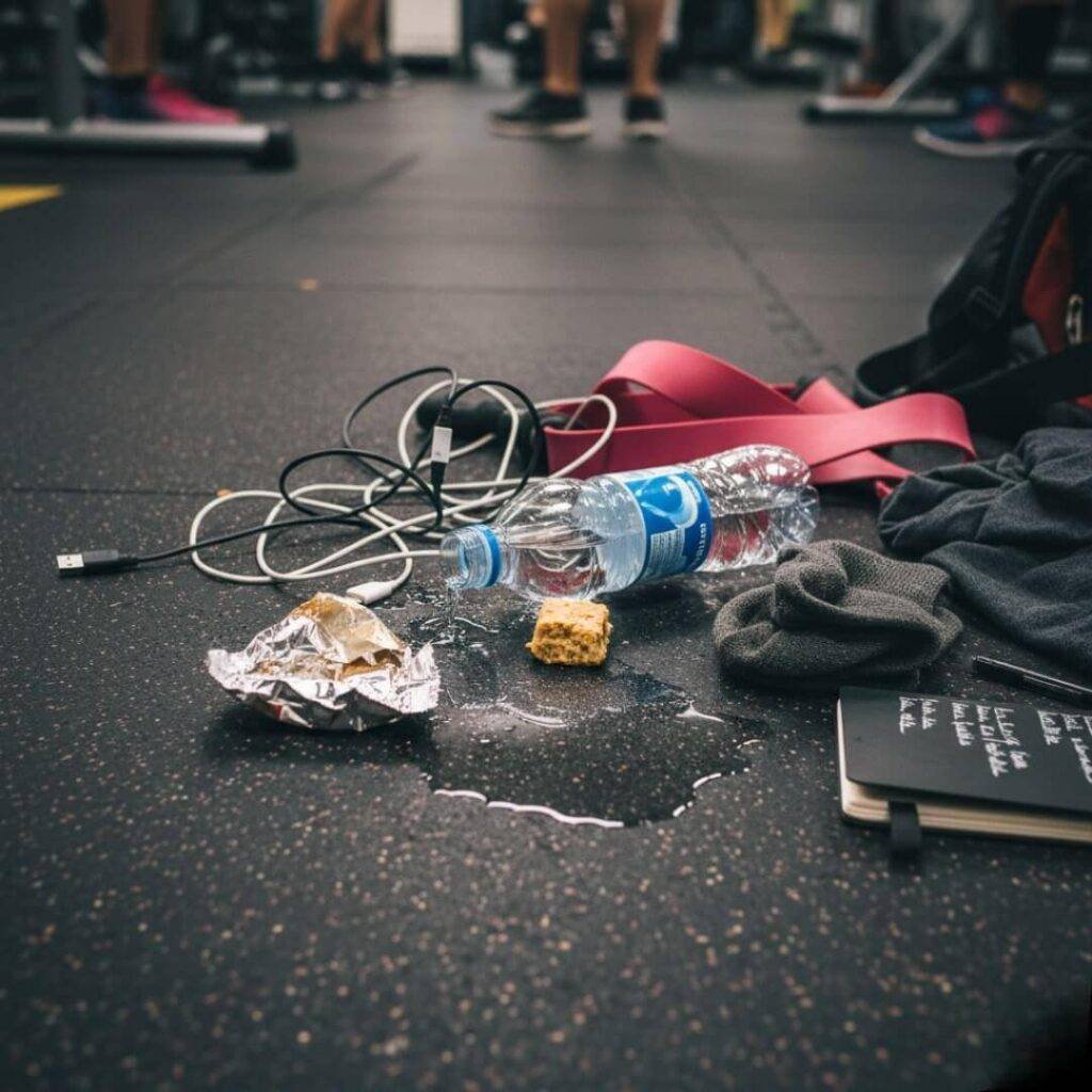 A low-angle, slightly blurred shot of a messy collection of gym bag contents scattered on a black gym floor. Visible items include a half-eaten granola bar on its wrapper, a leaky water bottle creating a small puddle, a tangled charging cord, a resistance band, and a small black notebook. The legs of other gym-goers are out of focus in the background.