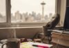 Daily Habits of Highly Successful People: Adopt Them Now! A cluttered desk with a steaming, chipped coffee mug, a planner with sticky notes, and a neon pink pen, all set against the blurry backdrop of a Seattle skyline with the Space Needle visible.